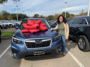 A blue Subaru Forester with one of those big, cheesy red bows on the hood and a smiling woman who just bought a car.