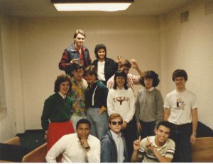 A group of somewhat nerdy freshmen in a dorm study lounge in the mid-80s. Of note is a girl with frizzy hair wearing a green sweater and a red skirt with a young Black man in front of her, attempting to strike a suave pose.