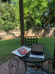 A laptop computer, a lap desk, and a red composition book sit on a metal mesh circular table in the foreground, with a green lawn, trees, and a wooden fence in the background.