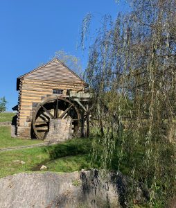 A grist mill made of logs and stones with a water wheel. There's a weeping willow tree in the foreground, and the sky is a bright blue.