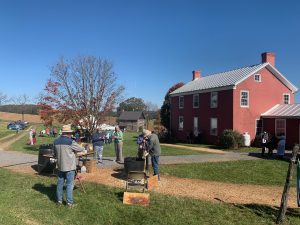 Demonstration day at a historic farm. In the foreground, a man makes popcorn in a big, black kettle over an open fire. To the right is a big, red-brick farmhouse. In the background is a white dining pavilion tent. The sky is a bright, clear, blue.