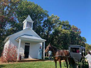 A white, wooden schoolhouse with a woman in an old-fashioned dress standing in front. In the foreground is an Amish-type black buggy pulled by a dark brown horse. There's a row of green trees behind the school, and the sky is bright blue.