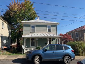 A two-story Victorian cottage with a front porch and ornate wood trim. The siding is a light grayish green. A blue Subaru is parked in front.