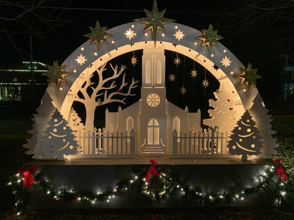 A Christmas display designed to look like cut paper, showing the silhouette of an old church building with an arch over it and white lights giving it a glow against the night sky.