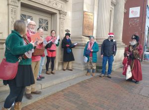 A group of people stand on the steps of an ornate early 20th century bank building, dressed for cold weather, holding music books. An old-fashioned Father Christmas in a fur hat stands with them.