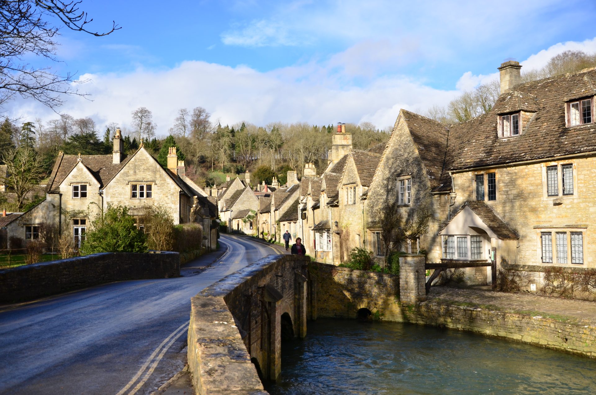 A brook runs under a stone bridge in the foreground. A lane crosses the bridge, passing between rows of golden stone cottages.