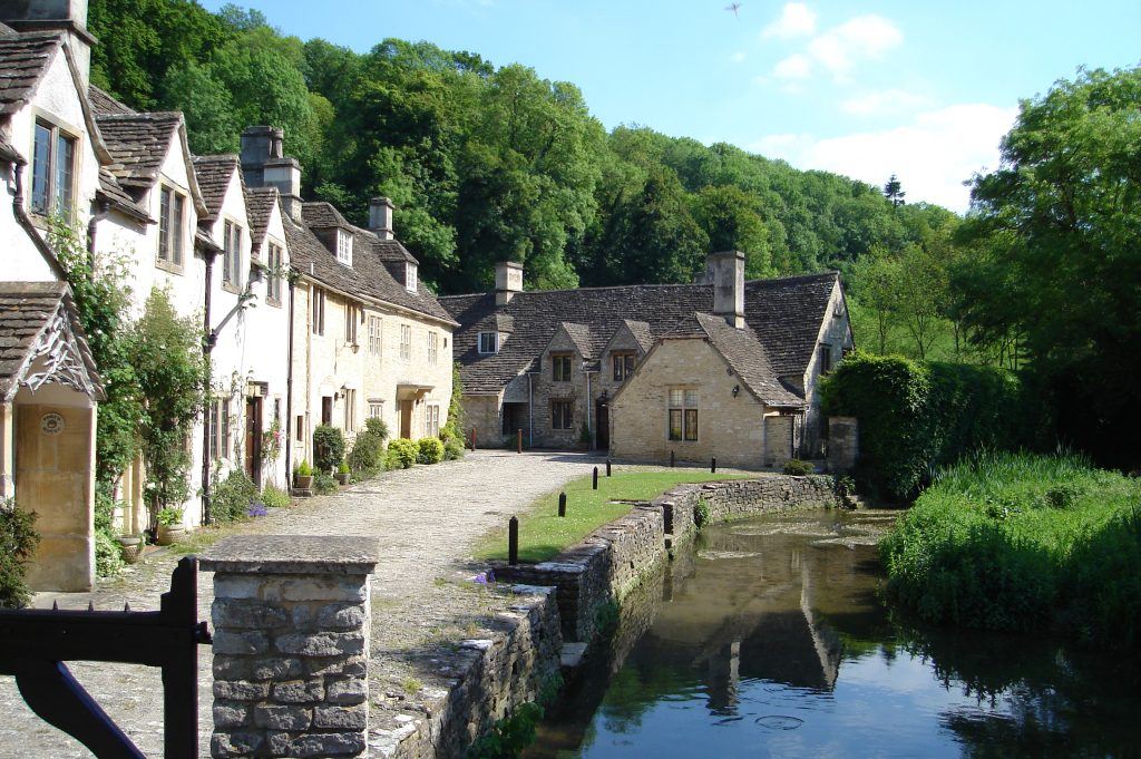 A row of buildings faces a brick-lined brook. A larger building with a steeply sloped slate roof stands facing the path that runs in front of the other buildings. There&#039;s a tree-covered hill in the background.