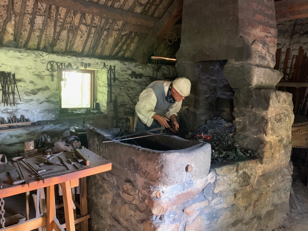 A stone hearth is in the middle of a forge guilding, with a pile of glowing coals in the middle and a chimney rising from behind it. To the left is a water trough with tools lying across it, and on the left of the photo is a workbench with tools and items made in the forge on display. A smith wearing 18th century costume digs in the coals with tongs.