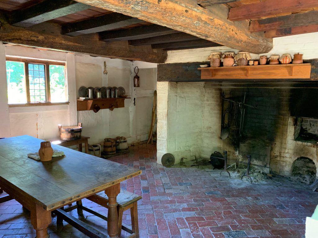 A kitchen in an old house, with red brick floor, large exposed beams in the ceiling, and a big fireplace, with racks for hanging pots over the fire. A wooden table with benchs sits to the left. There are shelves holding clay jars and pewter plates. A row of wooden buckets sits along one wall.