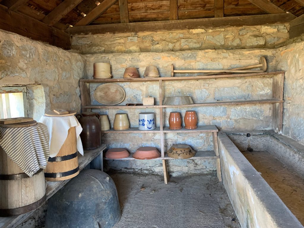 A frontier dairy, with stone walls, a high wooden roof, and a stone floor. There&#039;s a stone water trough on the right. At the rear are shelves holding clay jugs and bowls, as well as wooden butter churns. To the left on a shelf are barrels for churning butter, with cloths and wooden lids covering them.