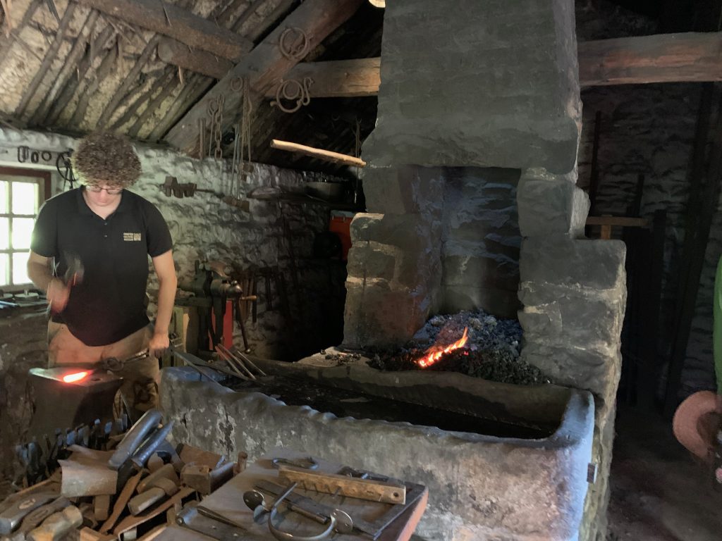 A smith pounds a piece of glowing metal laid on an anvil next to the hearth. Coals glow in the hearth.