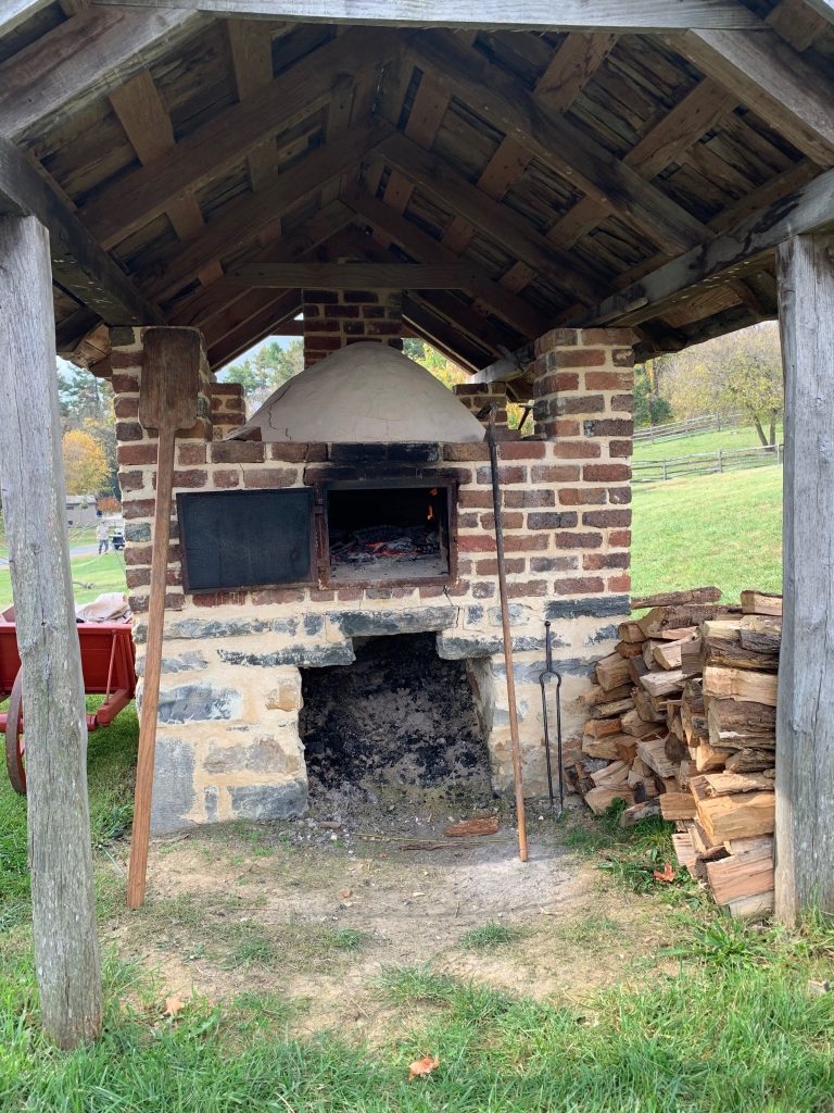 An outdoor brick oven, covered by a wooden shed. The door is open, showing glowing coals. There are ashes piled below the oven, and there&#039;s a stack of firewood to the right.