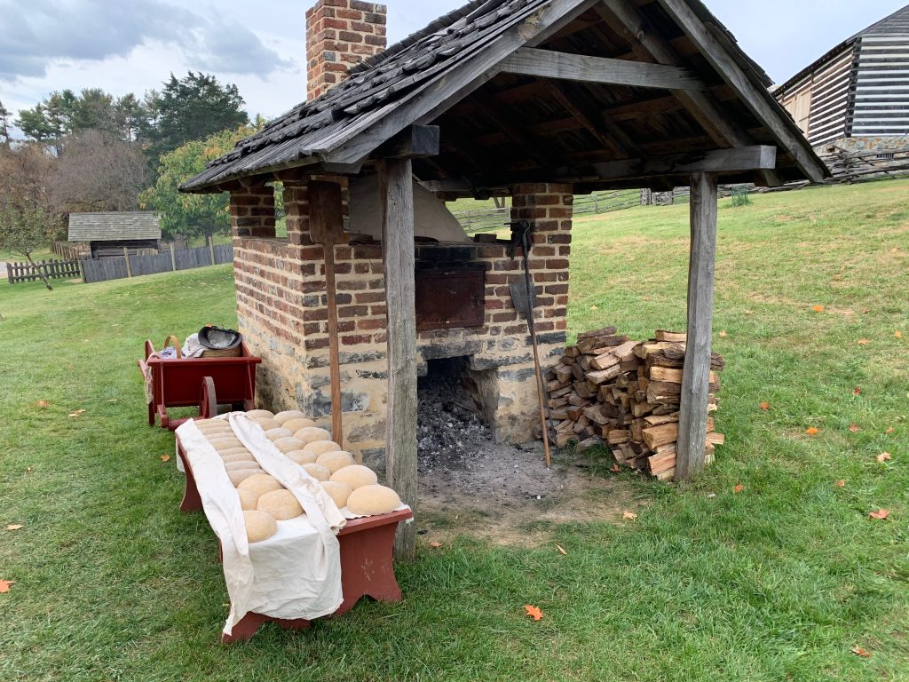 An outdoor brick oven. Cloth-draped benches stand next to it, covered with shaped loaves of bread dough.