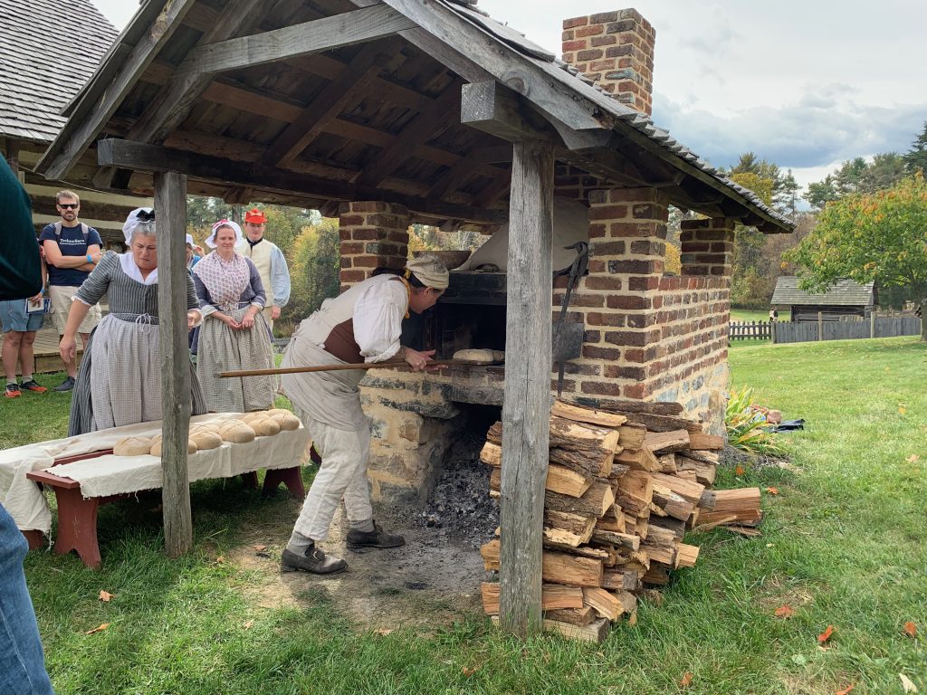 A baker in 18th century costume uses a wooden peel with a long handle to put loaves of bread into the brick oven. Women in costume stand nearby.