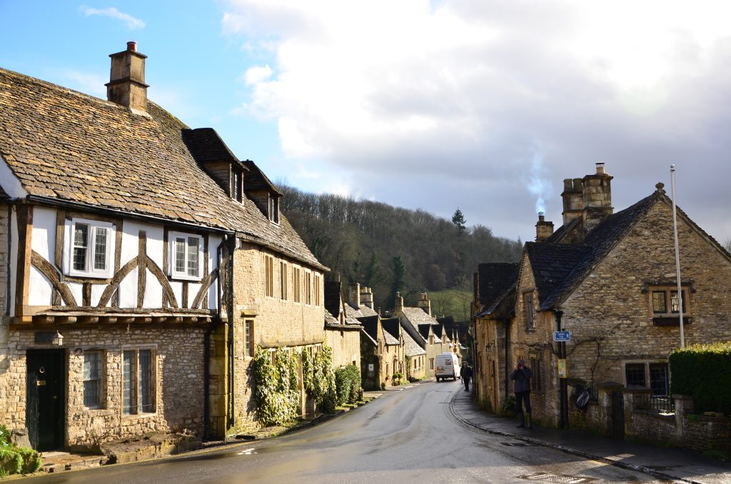 A village lane leads away from the market square, showing old stone buildings. The upper story of the building to the left is half-timbered. There&#039;s a tree-covered hill in the background.