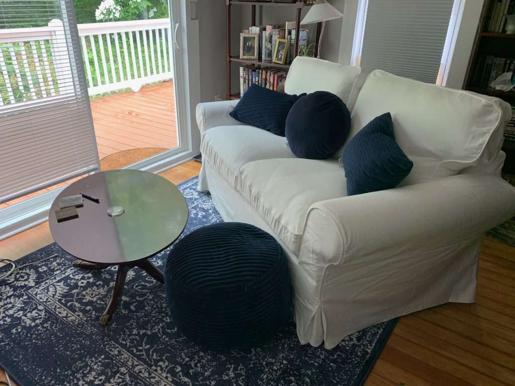A small den with old pine floors. There's a sliding glass door showing a view of a deck outside. A navy and white square rug lies on the floor. There's a small white sofa with navy blue throw pillows and a navy pouf footrest, and a dark wood oval coffee table in front of the sofa. In the background is a bookcase and a reading lamp. It's a cozy spot for spending a day reading.