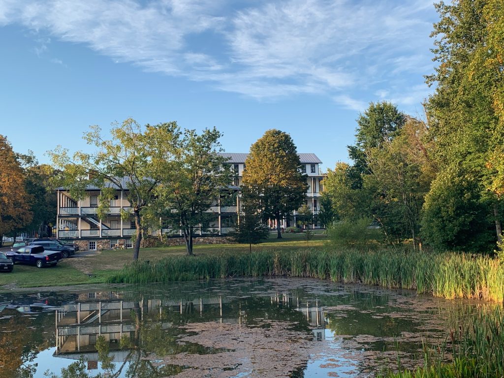 A Victorian hotel of white wood with green trim, and another larger one behind it. In the foreground is a small lake with the first building reflected in it.