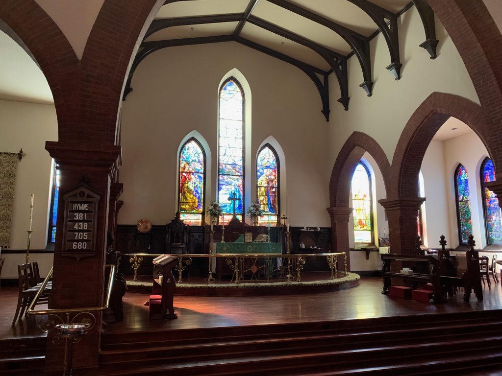 The interior of an old church building in Gothic Revival style. There are dark wood beams overhead, dark wood floors, pointed archways, and stained glass windows with light pouring in from outside.