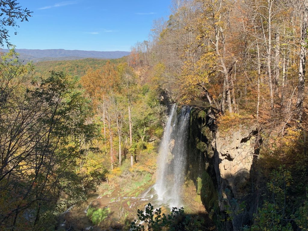 A waterfall spills off a hillside. It's surrounded by trees with faint autumn colors, and there's a blur of mountains in the background.