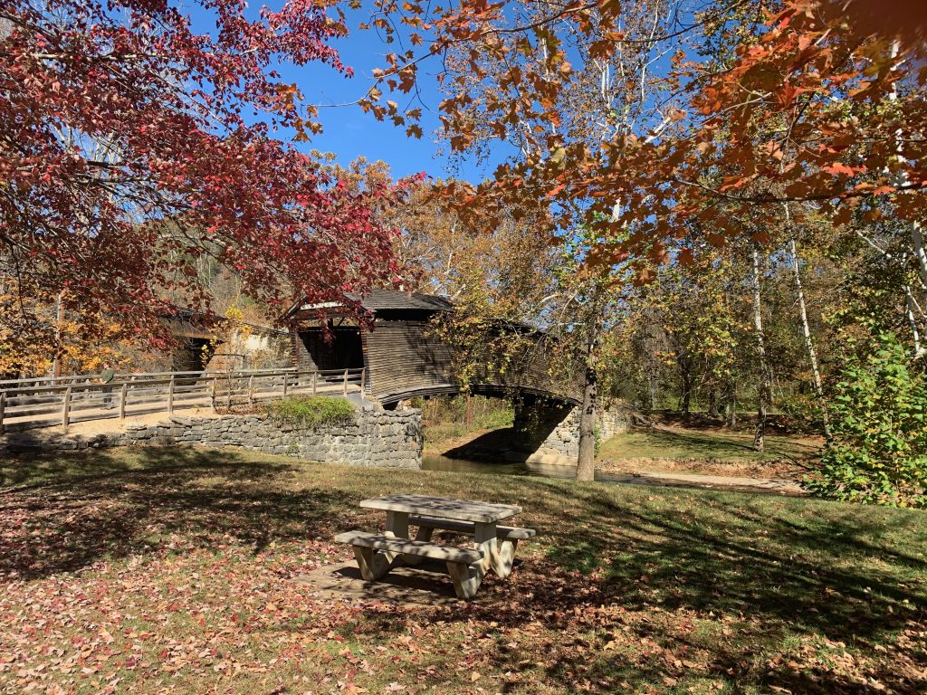 An old wooden covered bridge arches across a stream. In the foreground is a picnic table, and a tree with bright red leaves frames the picture.