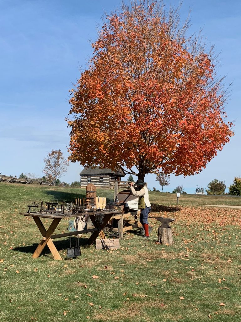 A blacksmith dressed in Revolutionary War-era attire works the bellows at an outdoor forge set under a tree blazing with bright orange leaves. Metal ware he's made are spread out on a nearby picnic table.