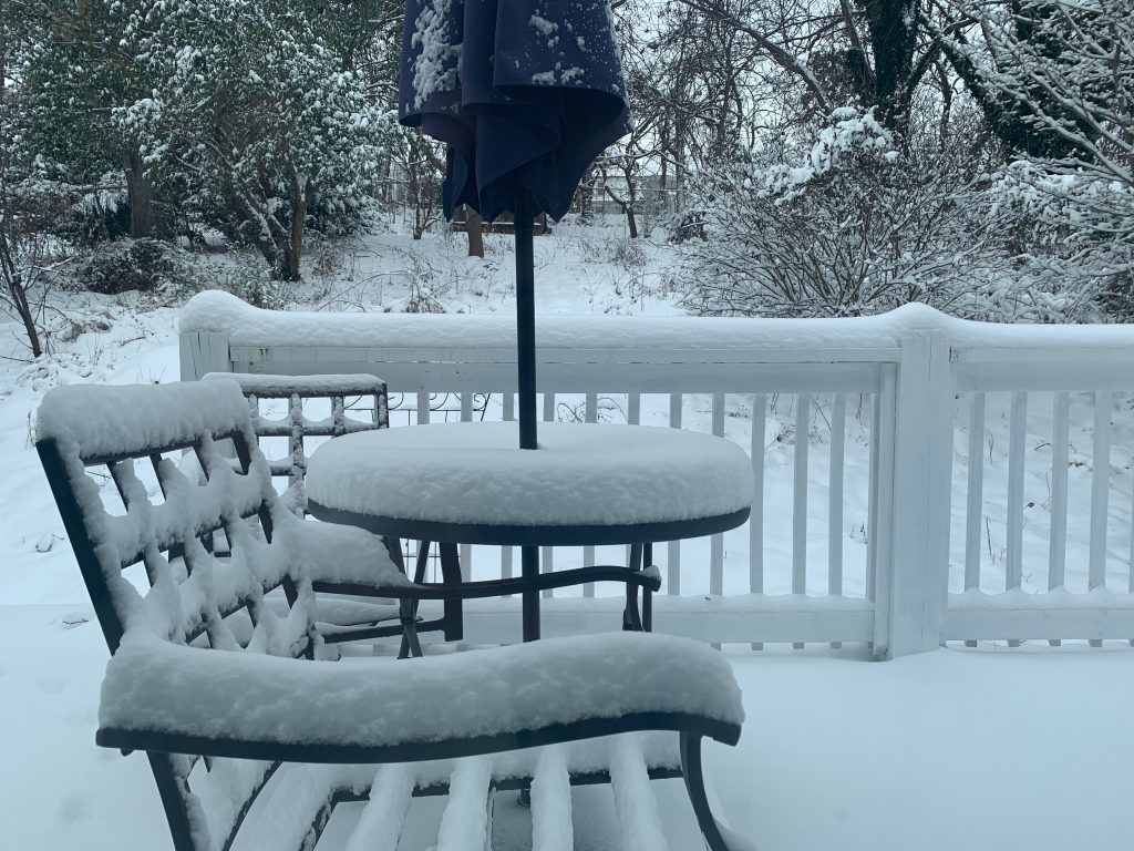 A thick layer of snow covers the top of a patio table, patio furniture, and deck railing.