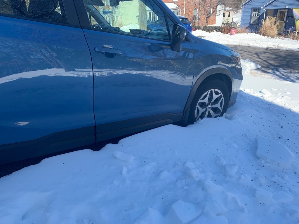 A blue Subaru Forester sits surrounded by what looks like fluffy snow but which is actually a thick crust of ice on top of snow. The snow comes up past the bottom of the doors.