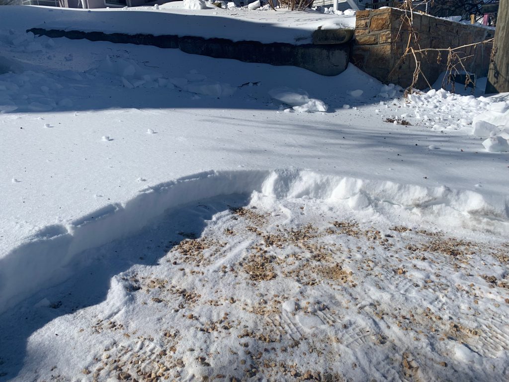 A cliff of ice shows a steep drop-off from the ice to the gravel driveway below.