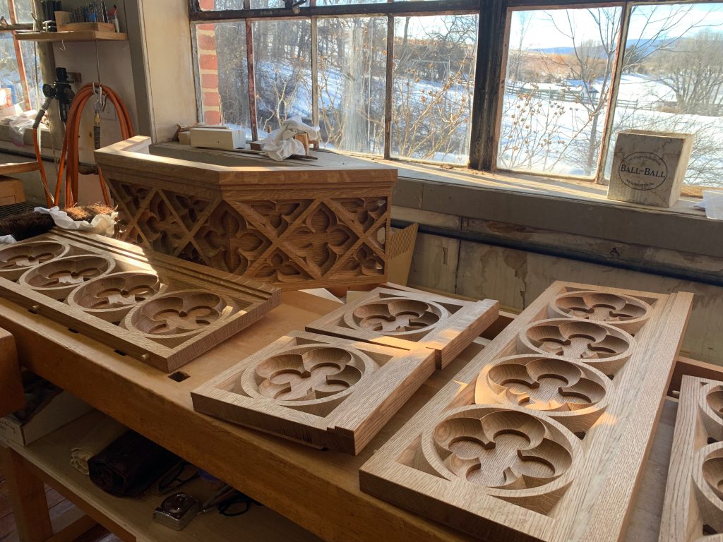 Bits of carved woodwork lie on a workbench. The view through the window behind is of snow-covered hills and mountains in the distance.