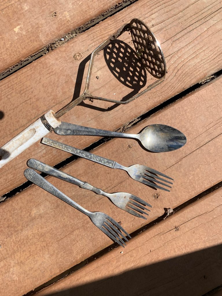 Several pieces of cutlery, including three forks and a soup spoon, plus a dirty, rusted potato masher.