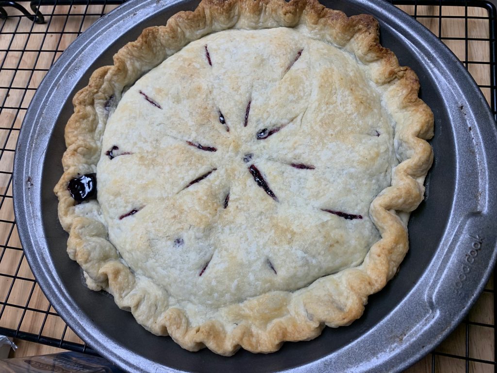 A light golden brown pie in a metal pie tin. The vents in the top crust are in a star-shaped pattern, and a bit of dark filling has oozed out of the crust in places.