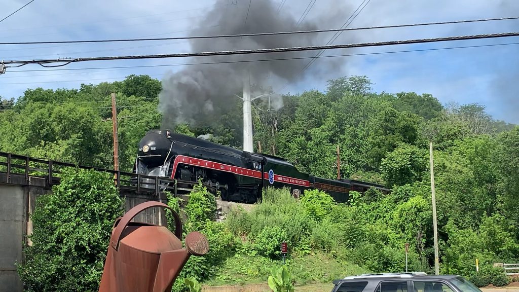 A sleek black steam train with red trim crosses a railroad trestle bridge, black smoke billowing from it. There's a large sculpture of a watering can in front of the bridge.