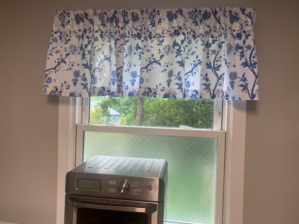 A kitchen window. A blue and white valance covers the top portion. The bottom has an etched-glass floral pattern. An air fryer oven sits in front of the window.