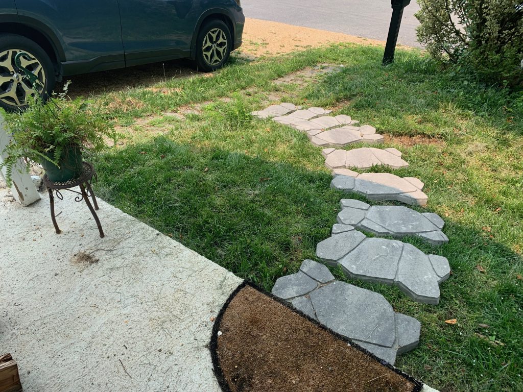 A path made of faux "natural" stepping stones leads from a front porch to a driveway. Along the edge of the driveway is a straight path of stones overgrown with weeds.