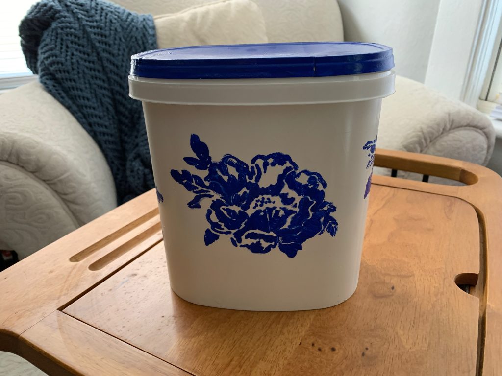 A repurposed dishwasher pod bin has a bright blue lid and a blue rose stenciled on the side.