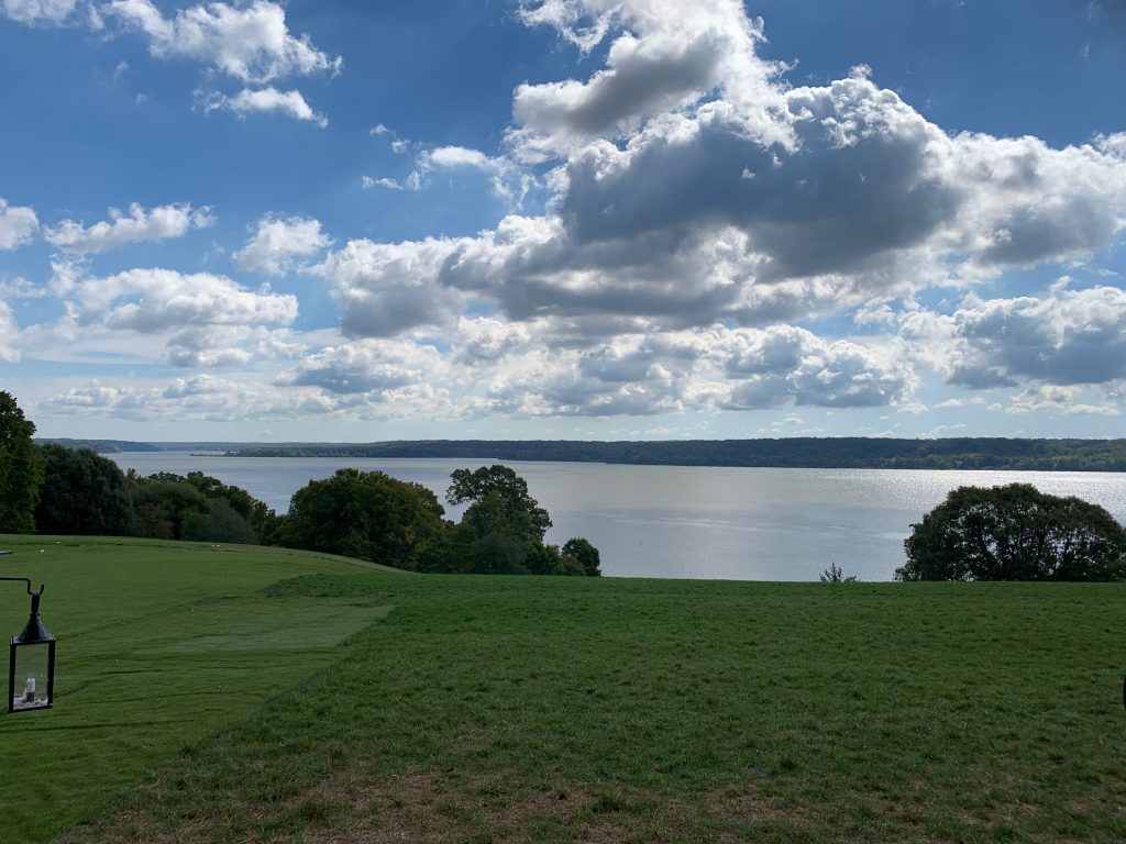 The view from the veranda at Mount Vernon. A grassy lawn slopes down to the river, with wooded land across the river. Puffy clouds dot a blue sky.